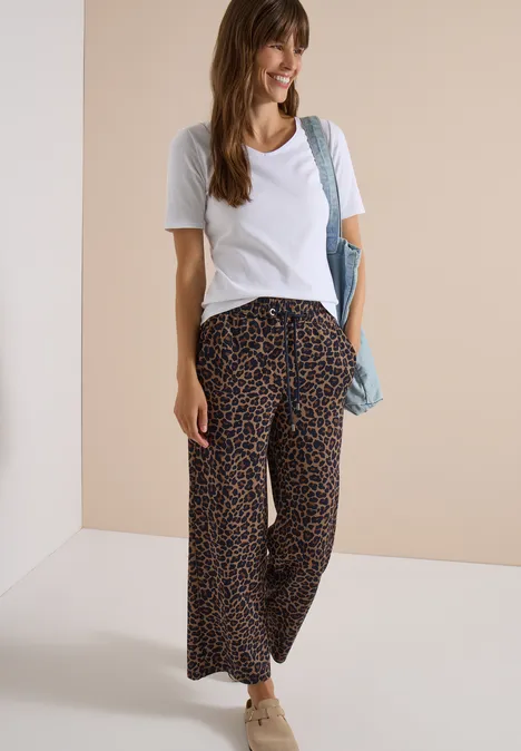 Femme souriante en t-shirt blanc, pantalon large léopard, baskets et sac d’épaule.
