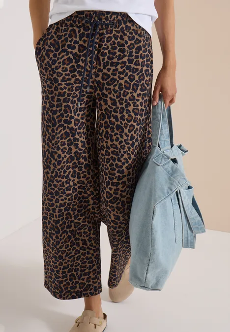 Une personne porte un pantalon léopard ample, des baskets claires et tient une veste en jean devant un fond neutre.