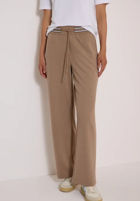 Weite hellbraune Cordhose mit Kordelzug, weiße Sneaker, Person vor weißem Hintergrund.