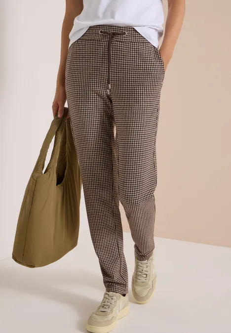 Eine Person trägt taillierte, karierte Hose, weiße Sneaker und hält eine große olivgrüne Tasche.