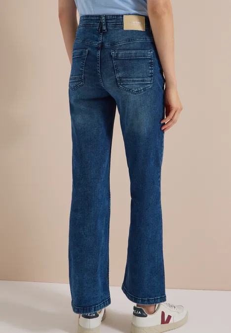 Frau steht von hinten, trägt dunkelblaue High-Waist-Jeans und weiße Sneaker vor beigeem Hintergrund.