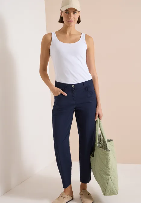 Frau in weißem Tanktop und dunkelblauer Hose trägt eine grüne Stepp-Tasche vor schlichtem Hintergrund.