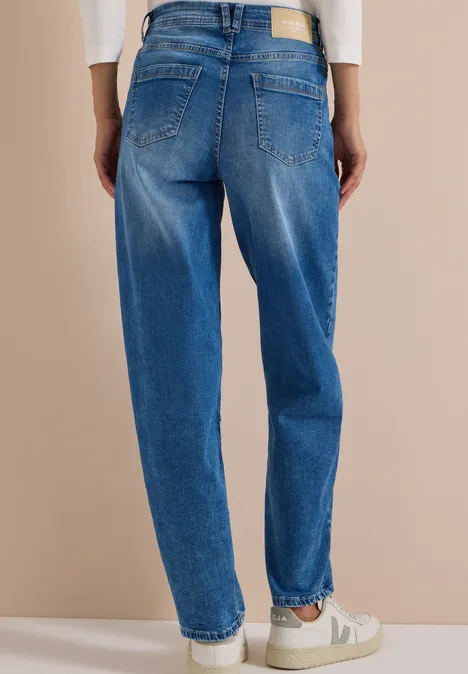 Vue de dos d’une personne portant un jean bleu ample et des baskets blanches sur fond beige.