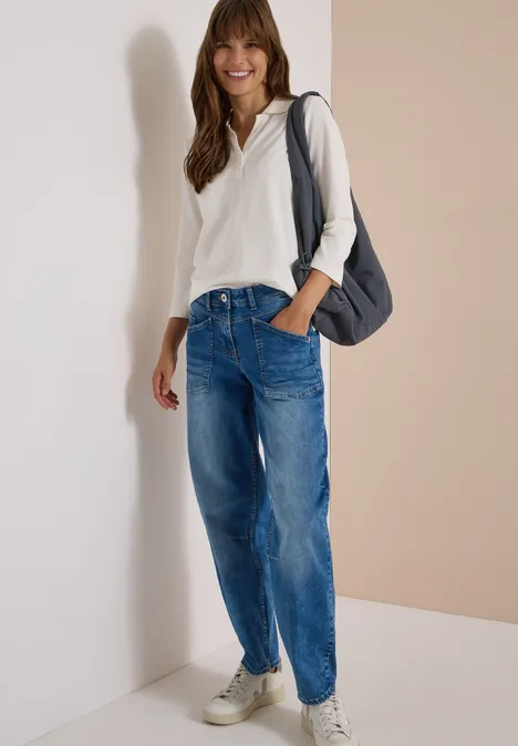 Femme en sweat à capuche blanc, jean ample et baskets, souriante, appuyée contre un mur, sac à bandoulière.