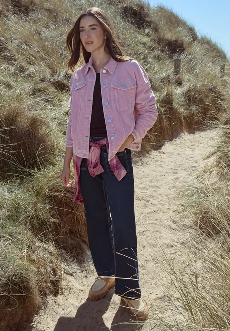 Frau in rosa Jeansjacke, weitem Dunkelblau-Jeans und beigen Sandalen läuft über einen grasigen Dünenpfad.