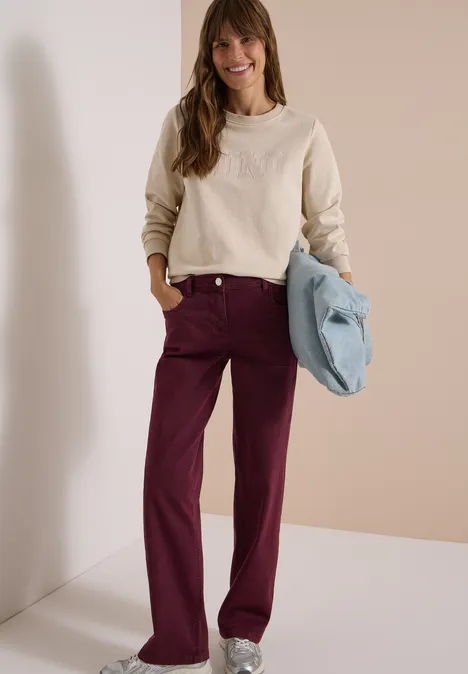 Femme debout sur fond neutre, porte un sweat beige, un pantalon bordeaux et tient un sac bleu clair.