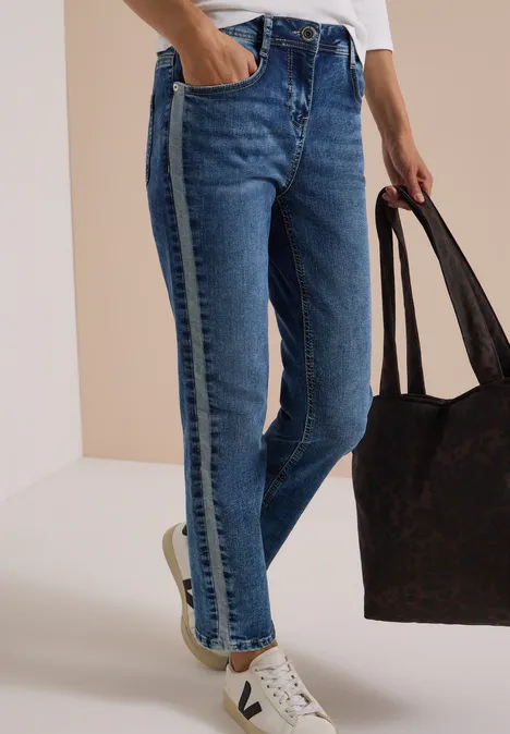 Une personne porte un jean bleu à bande latérale, des baskets blanches et tient un grand sac marron.