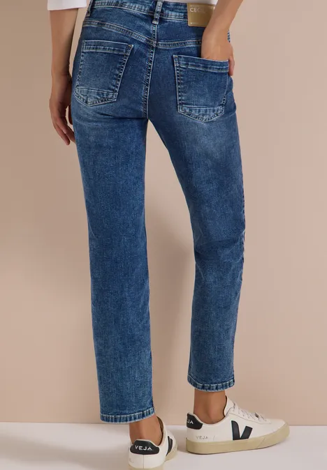 Femme en jean bleu taille haute, coupe droite, baskets blanches, sur fond beige.