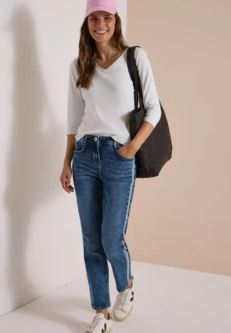 Femme souriante en top blanc, jean bleu, baskets blanches et casquette rose avec grand sac devant un mur neutre.