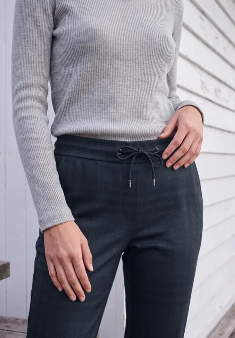 Frau in grauem Pulli und dunklen, taillierten Hosen mit Kordel vor weiß vertäfelter Wand.