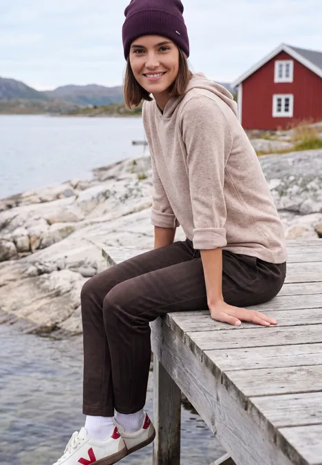 Eine lächelnde Frau in Mütze und beigem Pullover sitzt auf einem Holzsteg am See vor einer roten Hütte.