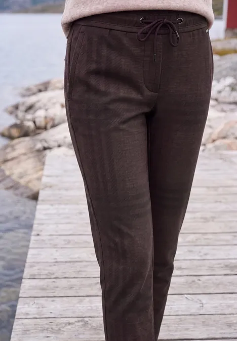 Eine Person steht auf einem Holzsteg und trägt eine braune Hose mit Kordelzug.