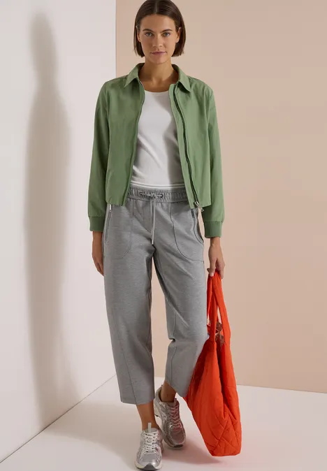 Frau in grüner Jacke, weißem T-Shirt und grauer Hose mit silbernen Schuhen hält eine rote Tasche.