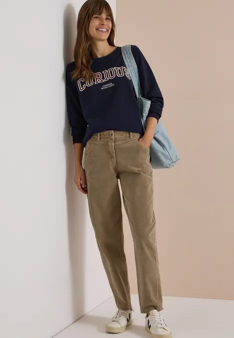 Frau lehnt an Wand, lächelt, trägt dunkelblaues Sweatshirt, olivgrüne Cordhose, weiße Sneaker und blaue Tasche.