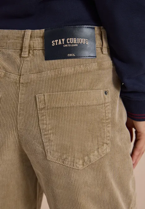 Beige Cordhose mit Gesäßtasche und schwarzem Label 'STAY CURIOUS', darüber dunkelblaues Oberteil.