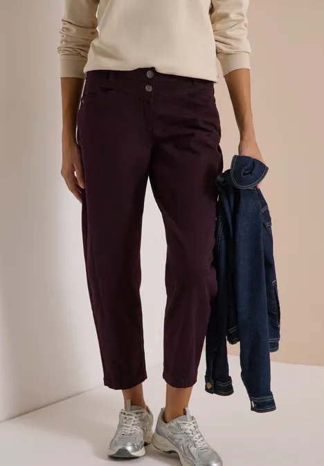 Person trägt beiges Oberteil, bordeauxrote Hose, silberne Sneaker und hält eine Jeansjacke.