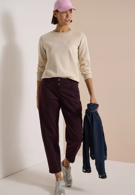 Frau trägt helles Langarmshirt, weinrote Hose, Baseballcap, silberne Sneaker und hält eine blaue Jacke.