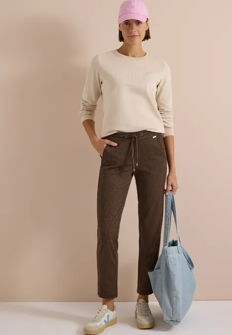 Frau mit rosa Cap, beigem Pullover, braunen Hosen und Sneakern, die eine hellblaue Tasche hält.