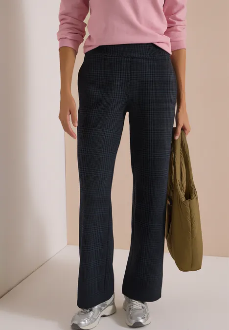 Frau trägt rosa Pullover, schwarze Hose und silberne Sneaker, hält einen grünen Beutel.