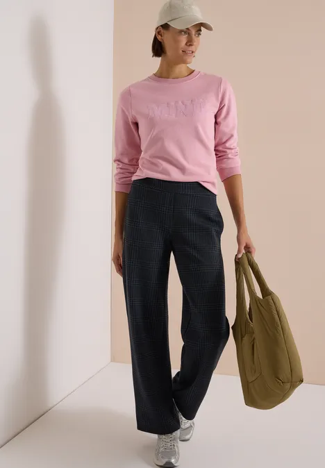 Frau in rosa Pullover, weiten dunkelblauen Hosen, weißer Cap und Sneakers, trägt olivgrüne Tasche.