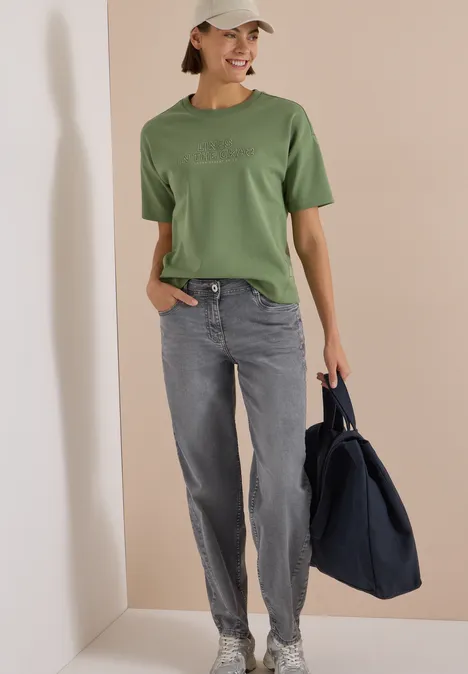 High Waist Barrel Leg Jeans w stylu Loose Fit light grey wash Kobieta w zielonym t-shircie, szarych dżinsach i czapce, trzyma dużą granatową torbę na beżowym tle.