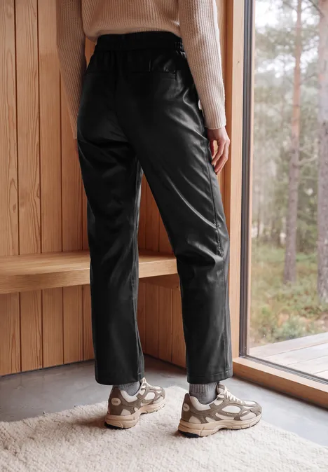 Eine Person trägt schwarze Lederhosen und Sneaker, steht vor einem Fenster mit Blick auf den Wald.