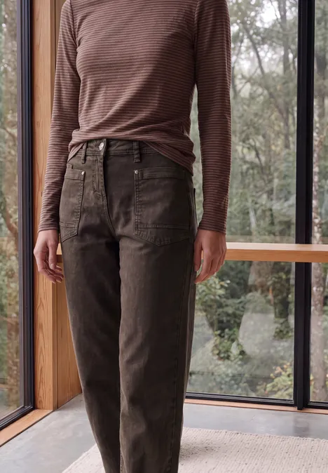 High Waist Barrel Leg Jeans im Loose Fit truffle brown Person steht vor großem Fenster, trägt braune Jeans und ein langärmliges Oberteil.