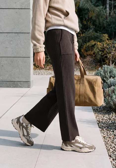 Personne marche sur un trottoir, pantalon plissé brun, baskets beiges et grand sac.
