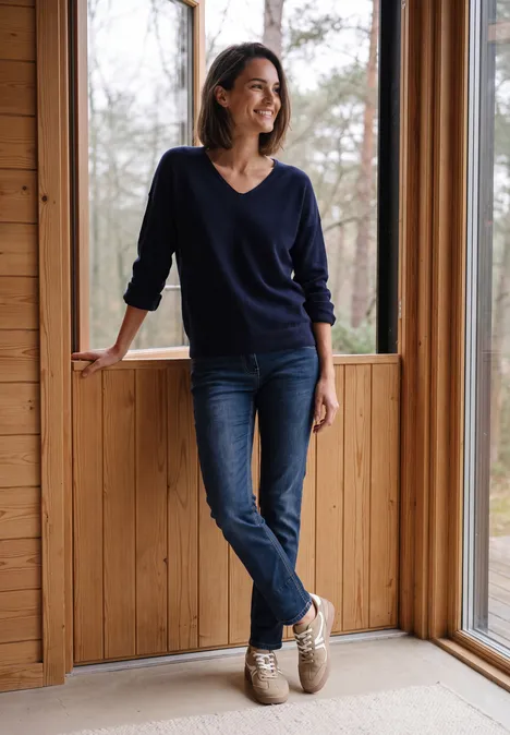 Eine Frau lehnt in einem Holzhaus an der Fensterrahmung, trägt Jeans und Pullover und lächelt entspannt.