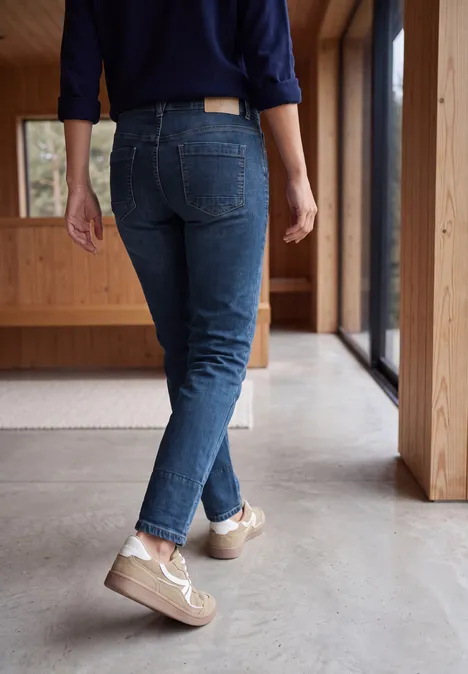 Rückenansicht einer Person in Jeans und hellen Sneakern auf einem modernen Betonboden nahe Fenstern.