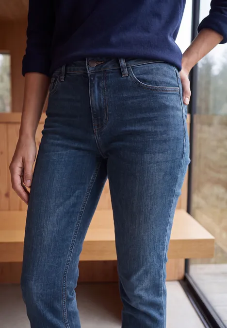 Person trägt hoch geschnittene dunkelblaue Jeans, steht neben einem Fenster vor Holzwand.