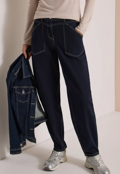 Frau trägt locker sitzende dunkelblaue Jeans, hält Jeansjacke, trägt silberne Sneaker.
