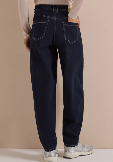 Person trägt dunkelblaue Jeans mit geradem Schnitt und weißen Sneakern, Rückansicht vor beigem Hintergrund.