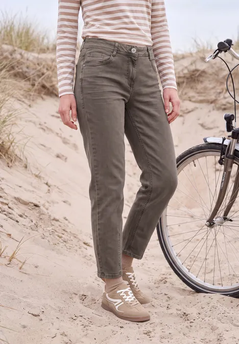 Persoon in olijfgroene broek, gestreepte trui en sneakers staat in zand naast een fiets.