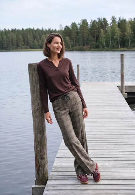 High Waist Wide Leg Hose im Loose Fit truffle brown Frau lehnt an Pfosten auf einem Steg, im Hintergrund See und Wald.