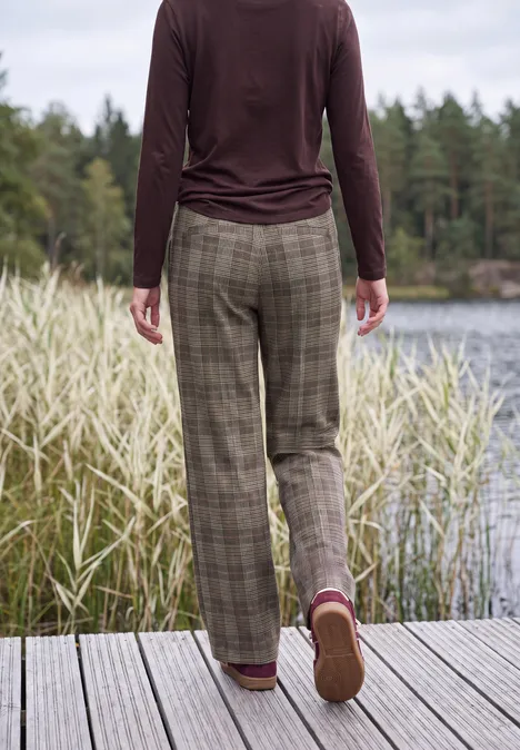 High Waist Wide Leg Hose im Loose Fit truffle brown Person in karierter Hose und Langarmshirt steht auf einem Steg am See, umgeben von Schilf und Wald.