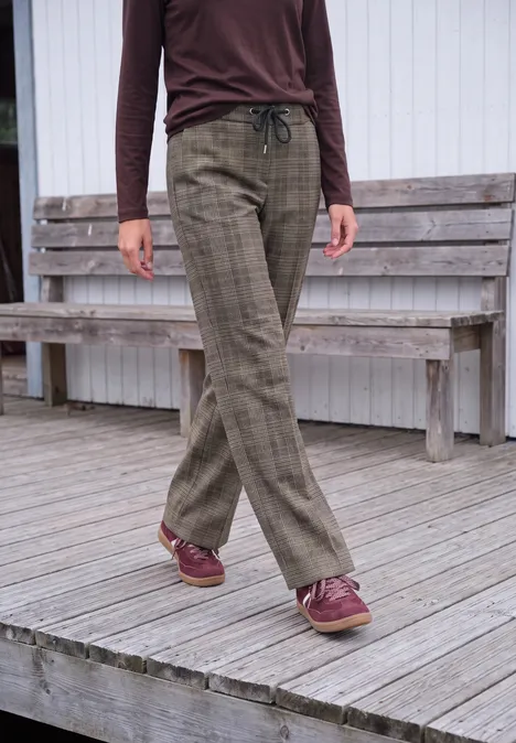 High Waist Wide Leg Hose im Loose Fit truffle brown Frau trägt braunes Oberteil, karierte, olivgrüne Hose und weinrote Schuhe auf Holzterrasse.