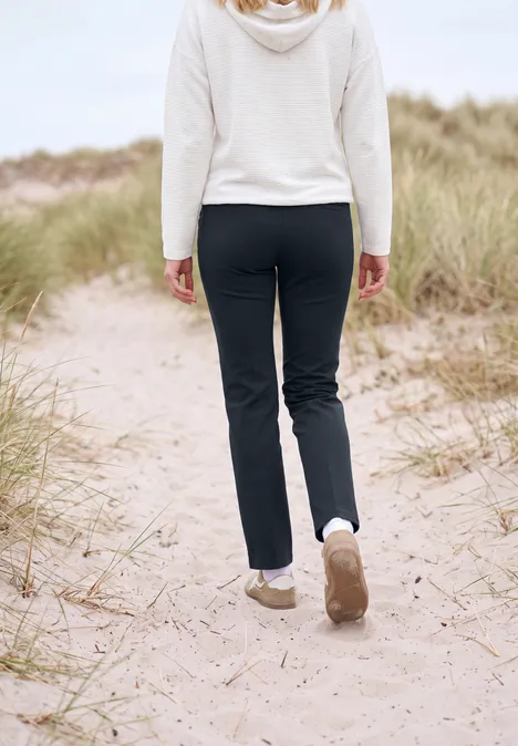 Une personne en pantalon noir et pull blanc marche sur un sentier de dunes sableuses avec herbes.