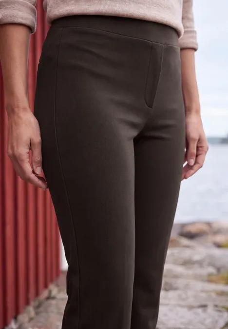 High Waist Bootcut Hose im Slim Fit truffle brown Frau steht an einem roten Holzhaus am Wasser und trägt dunkelgraue, eng anliegende Hose auf steinigem Weg.