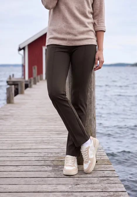 High Waist Bootcut Hose im Slim Fit truffle brown Person am Meer auf Holzsteg, trägt beigen Pullover, braune Hose und weiße Sneaker, rotes Bootshaus im Hintergrund.
