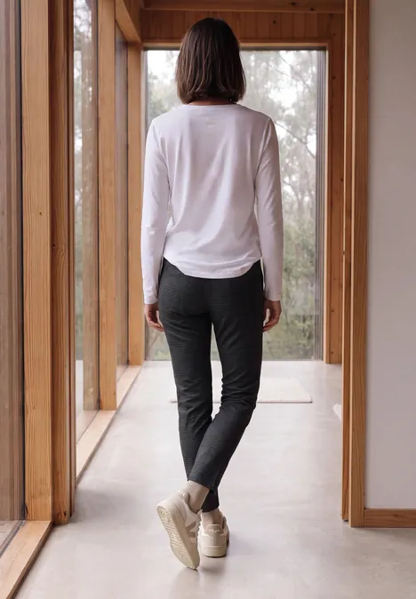 Mid Waist Slim Leg Hose im Casual Fit truffle brown Person steht im Gang mit Holzfenstern, Rücken zur Kamera, trägt weiße Bluse, schwarze Hose, helle Schuhe.
