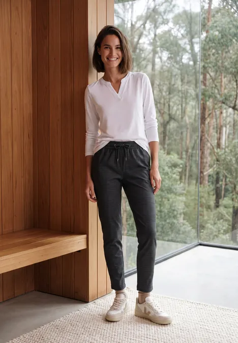 Mid Waist Slim Leg Hose im Casual Fit truffle brown Eine Frau steht in weißem Hemd und dunkler Hose lächelnd vor einem Fenster mit Waldblick.