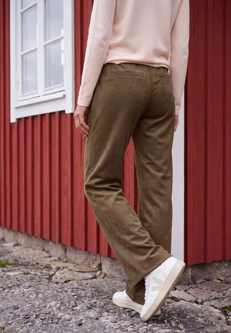 Person lehnt am roten Holzhäuschen, trägt olivegrüne Cordhose und weiße Sneaker.