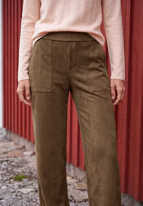 Damenhose aus grünem Cord mit großen Taschen, kombiniert mit rosafarbenem Langarmpullover vor roter Holzwand.