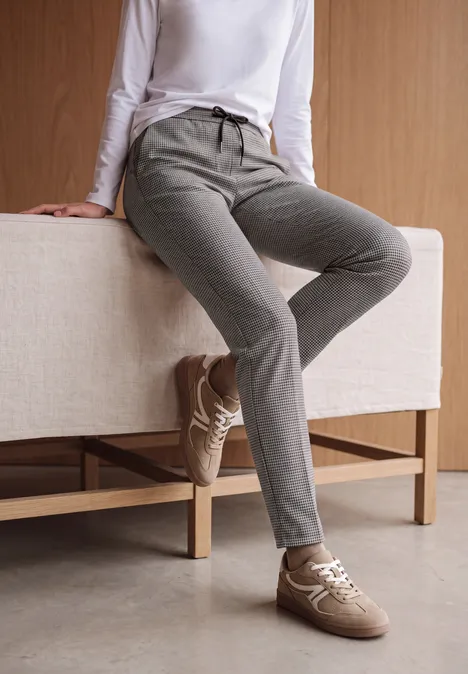 Slim Leg Hose mit Houndstooth-Muster cashmere beige Person sitzt entspannt auf einer Bank, trägt weißes Langarmshirt, graue Jogginghose und beige Sneaker.