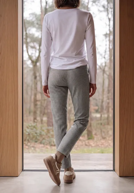 Slim Leg Hose mit Houndstooth-Muster cashmere beige Person in weißem Oberteil und grauer Jogginghose steht vor einem Fenster, blickt in den Wald