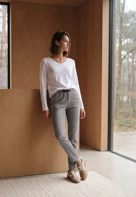 Slim Leg Hose mit Houndstooth-Muster cashmere beige Eine Frau in grauen Hosen und weißem Shirt lehnt in einem modernen, hellen Raum an einer Holzwand und schaut hinaus.