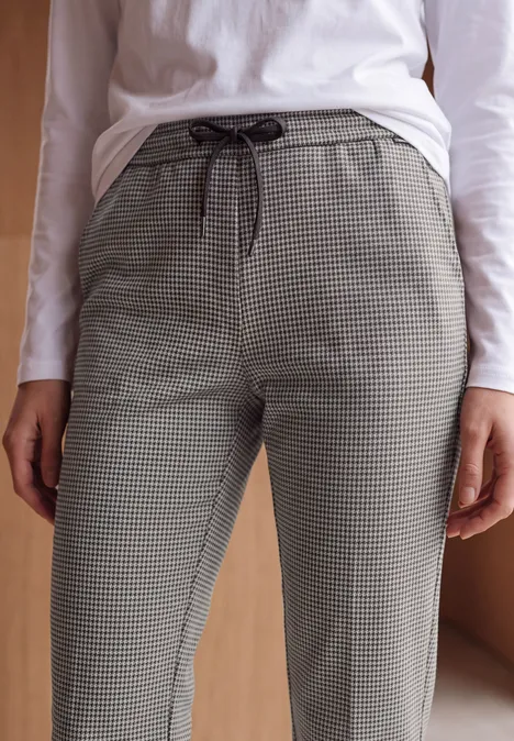 Slim Leg Hose mit Houndstooth-Muster cashmere beige Frau trägt graue Hahnentrittmuster-Hose und weißes Langarmshirt vor holzfarbenem Hintergrund.