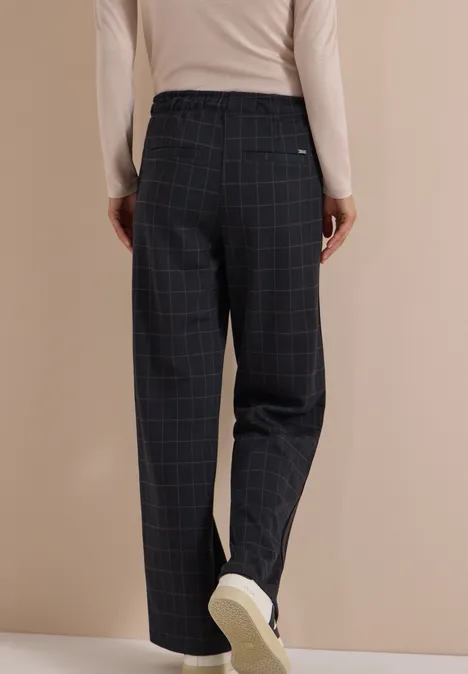 Wide Leg Hose mit Karomuster und Tape urban dark blue Person von hinten in locker sitzenden, dunkelkarierten Stoffhosen, beigem Oberteil und weißen Sneakern.
