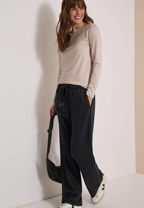 Wide Leg Hose mit Karomuster und Tape urban dark blue Frau trägt beiges Langarmshirt, schwarze Karohose, weiße Sneaker und schwarze Tasche, steht lächelnd.